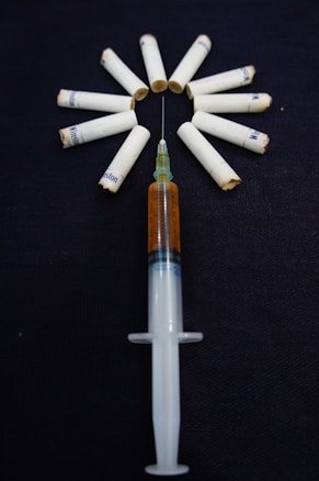 A syringe filled with a brown liquid is placed on a dark surface, surrounded by several cigarette filters arranged in a circle. The arrangement seems intentional and creates a striking visual with the cigarettes fanning out from the needle.