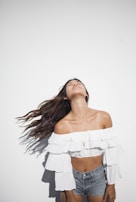 A person standing against a plain white background, wearing a white off-shoulder ruffled top and denim shorts. The person's head is tilted back with long hair flowing down, suggesting movement.