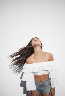 A person standing against a plain white background, wearing a white off-shoulder ruffled top and denim shorts. The person's head is tilted back with long hair flowing down, suggesting movement.