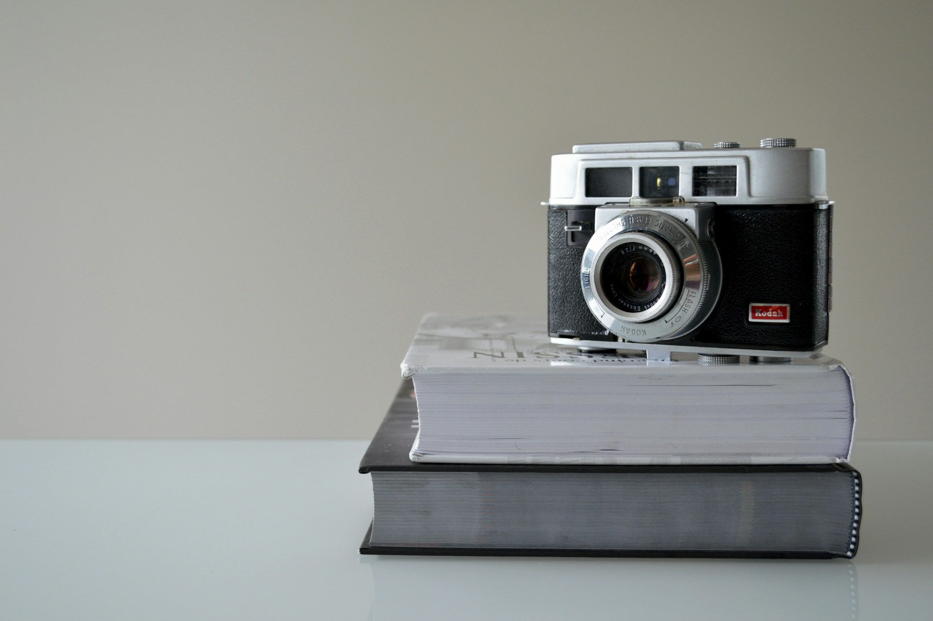A vintage camera is positioned on top of two closed hardcover books, resting on a clean, smooth surface. The background is a plain, light color, creating a minimalist and focused scene.