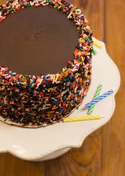 A chocolate cake topped with rainbow sprinkles, placed on a white cake stand with a scalloped edge. Several colorful candles, including blue, yellow, and green, are lying beside the cake on the stand. The background features a wooden tabletop.
