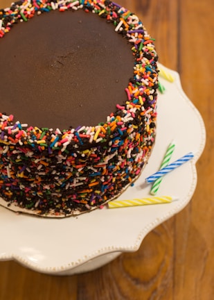 A chocolate cake topped with rainbow sprinkles, placed on a white cake stand with a scalloped edge. Several colorful candles, including blue, yellow, and green, are lying beside the cake on the stand. The background features a wooden tabletop.