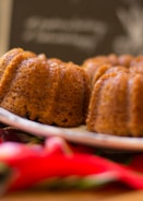 A close-up of freshly baked bundt cakes with a golden, textured crust. The cakes rest on a white plate, and vibrant red and green colors can be seen in the foreground, possibly a napkin. The background is blurred, suggesting focus on the cakes.