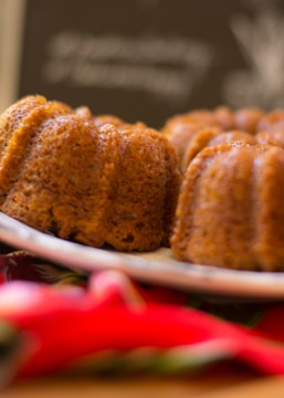 A close-up of freshly baked bundt cakes with a golden, textured crust. The cakes rest on a white plate, and vibrant red and green colors can be seen in the foreground, possibly a napkin. The background is blurred, suggesting focus on the cakes.