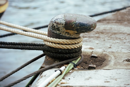 A weathered metal bollard with several ropes tied around it sits securely on a concrete dock. The ropes vary in color and thickness, including a light tan rope and a darker, almost black one. The surface of the bollard shows signs of rust and wear, indicating exposure to the elements. The background displays a blurred view of water, suggesting proximity to a harbor or waterfront.