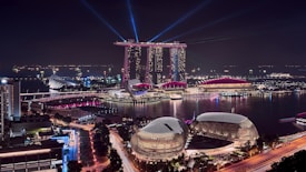 A vibrant night-time cityscape featuring a futuristic skyline with illuminated buildings and iconic structures. Multiple beams of light project into the sky, adding a dynamic quality to the scene. The reflection of lights on the water enhances the visual appeal, while city lights create a lively atmosphere.