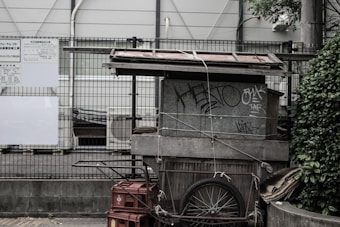 A makeshift structure is constructed using various materials including wood, metal sheets, and rope. It appears to be adjoined by a cart with a bicycle wheel, and some stacked crates. The structure is situated in an urban environment, with graffiti visible on its surface. Surrounding elements include a wire fence, a partially visible building, and some greenery.