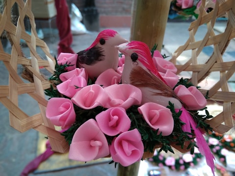 Two decorative birds with pink and brown feathers are nestled among synthetic pink roses on a woven base. The vivid pink flowers and birds create a vibrant and cheerful display.