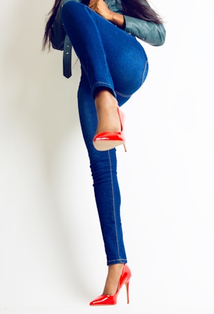 A person dressed in blue jeans and a dark jacket lifts their leg in a strong pose, showcasing bright red high-heeled shoes. The image conveys a sense of dynamic movement and bold fashion.
