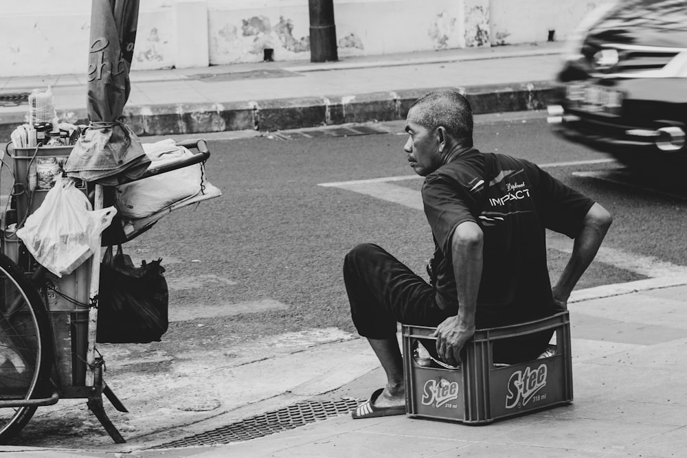 grayscale photo of man sits on crate beside road