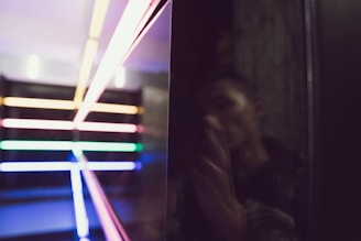 A series of brightly colored neon lights arranged horizontally and vertically create a geometric pattern against a dark background. On the right, a reflective surface shows a blurred image of a person holding a camera, adding an element of mystery.