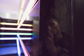 A series of brightly colored neon lights arranged horizontally and vertically create a geometric pattern against a dark background. On the right, a reflective surface shows a blurred image of a person holding a camera, adding an element of mystery.