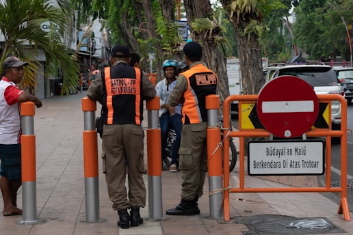 Two uniformed officers converse with pedestrians on a sidewalk. A pair of orange and black uniforms indicate they are members of a civil public order enforcement unit. A 'no entry' traffic sign and a warning sign in Indonesian is near them. Vehicles are visible on the adjacent street and a mix of palm trees and storefronts line the sidewalk.