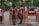 Two uniformed officers converse with pedestrians on a sidewalk. A pair of orange and black uniforms indicate they are members of a civil public order enforcement unit. A 'no entry' traffic sign and a warning sign in Indonesian is near them. Vehicles are visible on the adjacent street and a mix of palm trees and storefronts line the sidewalk.