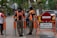 Two uniformed officers converse with pedestrians on a sidewalk. A pair of orange and black uniforms indicate they are members of a civil public order enforcement unit. A 'no entry' traffic sign and a warning sign in Indonesian is near them. Vehicles are visible on the adjacent street and a mix of palm trees and storefronts line the sidewalk.