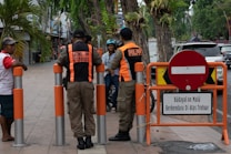Two uniformed officers converse with pedestrians on a sidewalk. A pair of orange and black uniforms indicate they are members of a civil public order enforcement unit. A 'no entry' traffic sign and a warning sign in Indonesian is near them. Vehicles are visible on the adjacent street and a mix of palm trees and storefronts line the sidewalk.