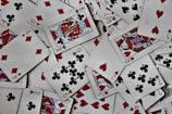 A scattered arrangement of playing cards featuring various suits and numbers. The cards include spades, diamonds, hearts, and clubs, with both face cards and numbered cards visible. The scene is chaotic, with cards overlapping each other in a random fashion.