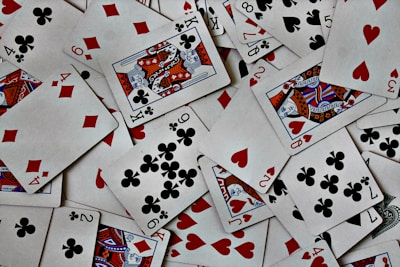 A scattered arrangement of playing cards featuring various suits and numbers. The cards include spades, diamonds, hearts, and clubs, with both face cards and numbered cards visible. The scene is chaotic, with cards overlapping each other in a random fashion.