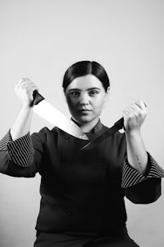 A person wearing a chef's uniform is holding two large kitchen knives, crossing them in front of their torso. The image is in black and white, emphasizing shadows and contours.