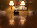 A warmly lit dining room features a long wooden table with a glossy finish. Two elegant lamps on either side cast a soft glow, illuminating a decorative floral painting on the wall. The room gives off a cozy and intimate atmosphere, framed by lightly colored chairs.