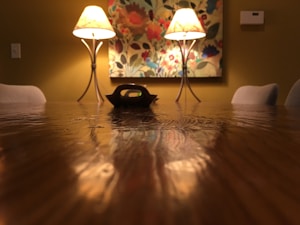 A warmly lit dining room features a long wooden table with a glossy finish. Two elegant lamps on either side cast a soft glow, illuminating a decorative floral painting on the wall. The room gives off a cozy and intimate atmosphere, framed by lightly colored chairs.