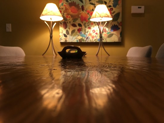 A warmly lit dining room features a long wooden table with a glossy finish. Two elegant lamps on either side cast a soft glow, illuminating a decorative floral painting on the wall. The room gives off a cozy and intimate atmosphere, framed by lightly colored chairs.