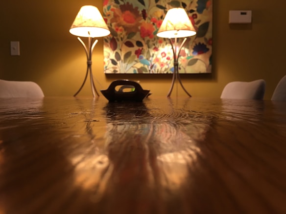 A warmly lit dining room features a long wooden table with a glossy finish. Two elegant lamps on either side cast a soft glow, illuminating a decorative floral painting on the wall. The room gives off a cozy and intimate atmosphere, framed by lightly colored chairs.