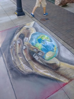 A chalk drawing on the pavement depicts a large, realistic hand cradling a small Earth. The artwork is detailed, with the continents visible on the globe. Above the artwork, a person wearing shorts and blue sneakers is walking on the sidewalk.