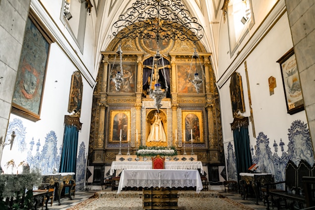 An ornate church altar featuring religious artwork and a central statue, surrounded by intricate gold detailing. Elaborate chandeliers hang from the ceiling, and the walls display decorative paintings and blue-patterned tiles. A white cloth covers the altar table, adorned with floral arrangements.