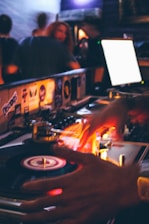 A DJ is skillfully working with a turntable, with hands in motion over the spinning vinyl records. Bright, colorful lights illuminate the scene, creating a vibrant and lively atmosphere. In the background, people are seen enjoying a night out in a dimly lit club or bar setting.