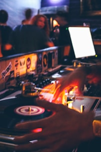 A DJ is skillfully working with a turntable, with hands in motion over the spinning vinyl records. Bright, colorful lights illuminate the scene, creating a vibrant and lively atmosphere. In the background, people are seen enjoying a night out in a dimly lit club or bar setting.