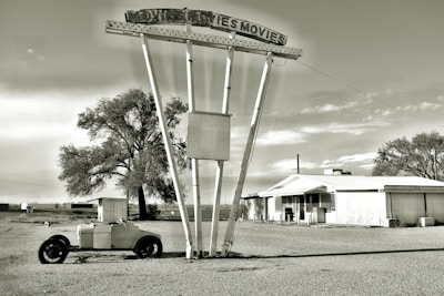 An abandoned drive-in movie theater with a large, deteriorating metal sign reading 'MOVIES MOVIES'. In front of the sign, an old, rusty trailer sits on a gravel lot. A small, run-down building is visible to the right, and there are sparse trees and clear skies in the background, conveying a sense of desolation and nostalgia.