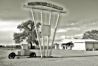 An abandoned drive-in movie theater with a large, deteriorating metal sign reading 'MOVIES MOVIES'. In front of the sign, an old, rusty trailer sits on a gravel lot. A small, run-down building is visible to the right, and there are sparse trees and clear skies in the background, conveying a sense of desolation and nostalgia.