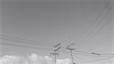 Multiple power lines are suspended on utility poles against a backdrop of scattered clouds in the sky. The poles are supporting various cables and wires extending in different directions.