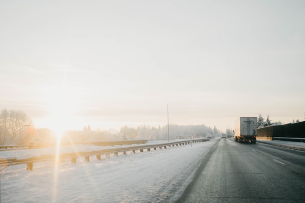 A winter highway scene with a large truck driving along a snow-covered road. The sun is low on the horizon, casting a warm glow and long shadows. Sparse trees line the road, and the sky is clear with soft hues.