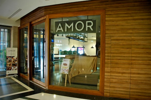 A storefront with large glass windows and a wooden frame, displaying the word 'AMOR' prominently above the entrance. Inside, a few light fixtures are visible hanging from the ceiling. There's a poster stand with images of baked goods near the entrance. The surrounding area has a clean, modern interior design with a polished black and white tiled floor.