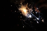 A close-up of a sparker being held by a hand, emitting bright sparks and trails of smoke against a dark background. The light from the sparker creates dynamic, bright lines and bursts, adding contrast to the darkness surrounding it.