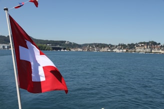 An image of a red and white flag flying on a pole near a large body of water, with a view of a distant town along the shoreline. The skyline includes various buildings and a backdrop of green hills.