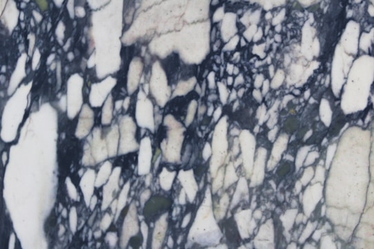 A marble surface displaying intricate black and white veining patterns with a few subtle hints of green. The design features irregular, jagged shapes interwoven amidst a backdrop of creamy white.