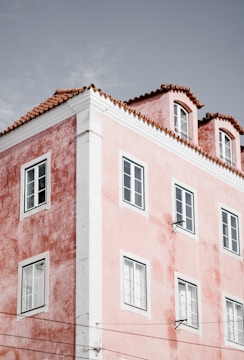 A pink stucco building with white trim and several windows, featuring a red-tiled roof against a light gray sky.