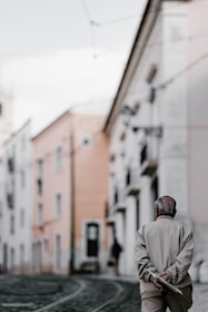 An elderly person walks down a quiet, narrow street with tram tracks. The buildings on either side are a mix of pastel colors with a slightly worn appearance, giving a sense of nostalgia. The person's posture is relaxed, conveying a sense of calmness or reflection.