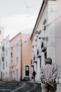 An elderly person walks down a quiet, narrow street with tram tracks. The buildings on either side are a mix of pastel colors with a slightly worn appearance, giving a sense of nostalgia. The person's posture is relaxed, conveying a sense of calmness or reflection.