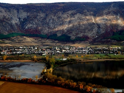 A small town is nestled at the base of a large, rocky mountain. The landscape includes a body of water reflecting the mountain's features, fields with autumn foliage, and scattered houses with a church steeple visible. The mountain is covered with patches of greenery and rock, creating a striking contrast.