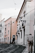 An elderly person walks alone down a narrow, cobblestone street lined with pastel-colored buildings. Overhead, wires from a tram system are visible. The street is quiet and empty, with the person appearing thoughtful and reflective.