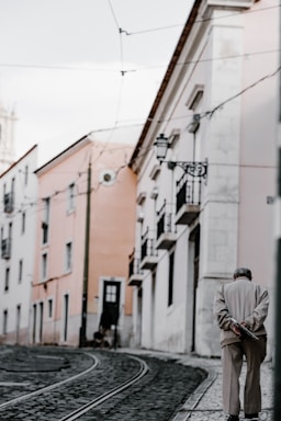 An elderly person walks alone down a narrow, cobblestone street lined with pastel-colored buildings. Overhead, wires from a tram system are visible. The street is quiet and empty, with the person appearing thoughtful and reflective.