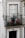A balcony adorned with intricate wrought iron railing holds potted plants with pink flowers. The exterior wall features a decorative tile pattern with floral designs in black, white, and ochre tones. A traditional white-framed window with lace curtains is above a black door numbered 16.