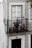 A balcony adorned with intricate wrought iron railing holds potted plants with pink flowers. The exterior wall features a decorative tile pattern with floral designs in black, white, and ochre tones. A traditional white-framed window with lace curtains is above a black door numbered 16.