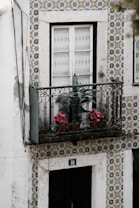 A balcony adorned with intricate wrought iron railing holds potted plants with pink flowers. The exterior wall features a decorative tile pattern with floral designs in black, white, and ochre tones. A traditional white-framed window with lace curtains is above a black door numbered 16.