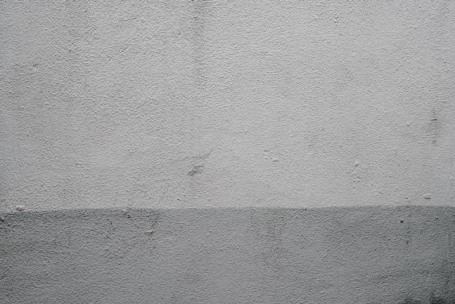 A textured concrete wall displaying a subtle two-tone gradient. The top part of the wall is a light gray while the bottom is a slightly darker shade. Minimalistic in style, the surface features small imperfections and marks.