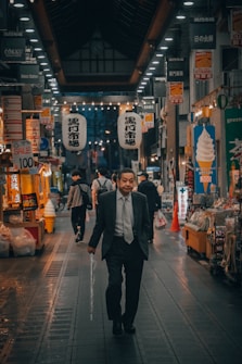 A man in a suit walks through a busy indoor market. The market is well-lit with numerous hanging signs and paper lanterns featuring Japanese characters. Various stalls display goods for sale under the yellow-orange glow of lights. Shoppers and pedestrians are seen in the background, creating a lively urban atmosphere.